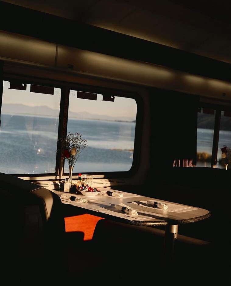 Dining table set for a meal in a train car with a view of a lake and mountains, bathed in warm sunlight and casting shadows.