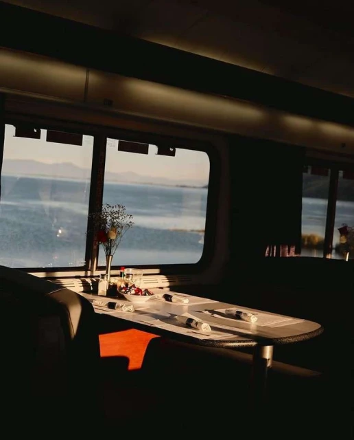 Dining table set for a meal in a train car with a view of a lake and mountains, bathed in warm sunlight and casting shadows.