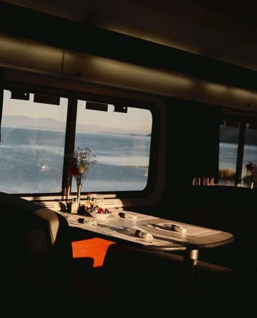 Dining table set for a meal in a train car with a view of a lake and mountains, bathed in warm sunlight and casting shadows.