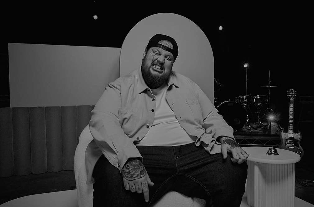 Monochrome portrait of a tattooed man seated in a modern chair, singing with his eyes closed, set against a music performance backdrop.