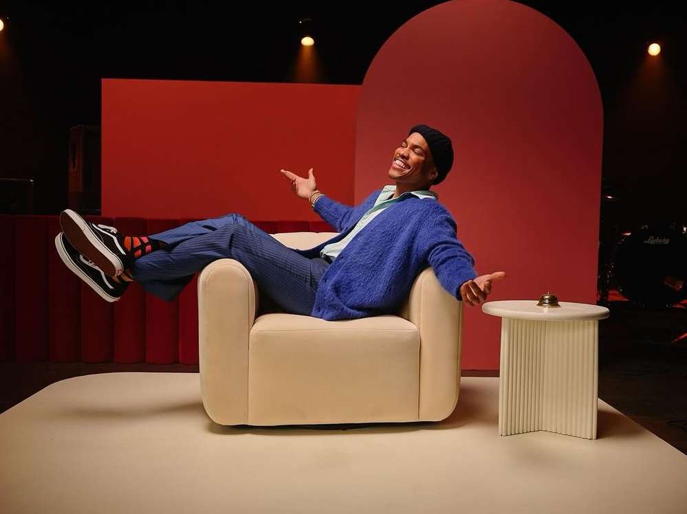 Man in blue cardigan and striped pants lounges in cream chair with Vans sneakers in front of red backdrop, black drum kit.