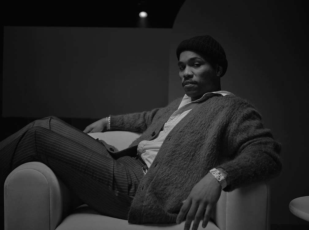 Stylish African American man in a buttoned cardigan and striped pants poses in a white chair against a grey background.