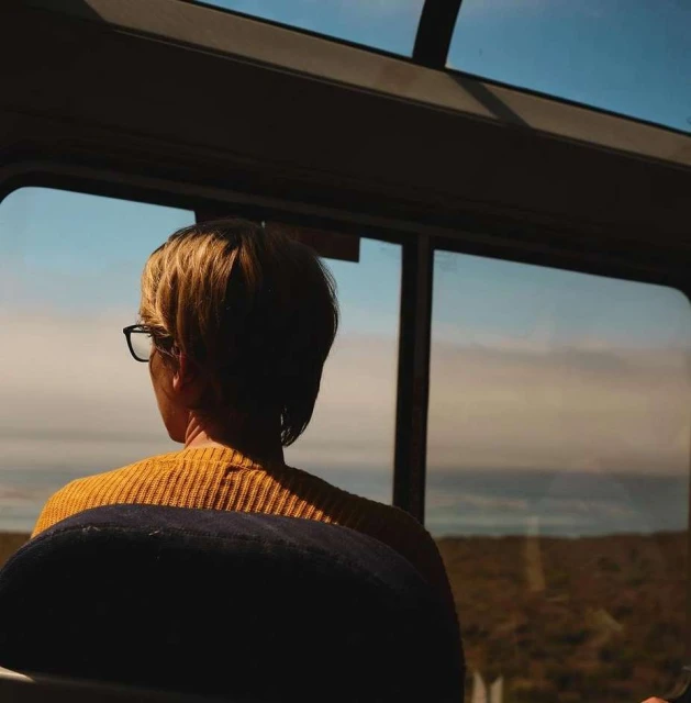 Woman in mustard sweater gazes out train window at ocean view. Back of head shows short blonde hair and black glasses.
