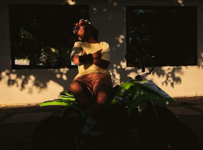 Child sits on a green toy quad bike in a sunlit outdoor setting with shadows on a white wall, wearing sunglasses and a yellow shirt.