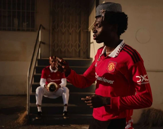 Two men in sports jerseys pose outdoors near stairs; one holds a soccer ball in an urban setting with neutral tones and a streetwear style.