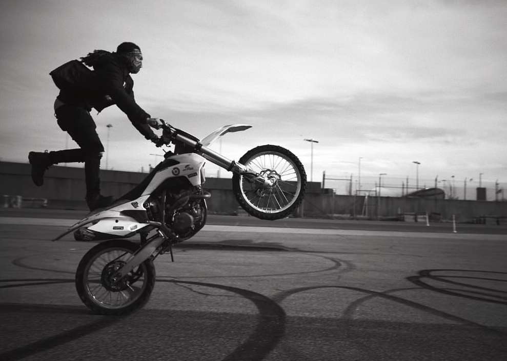 Motorcyclist performs a wheelie on a dirt bike in black and white, showcasing skill and balance with a dynamic urban backdrop and cloudy sky.