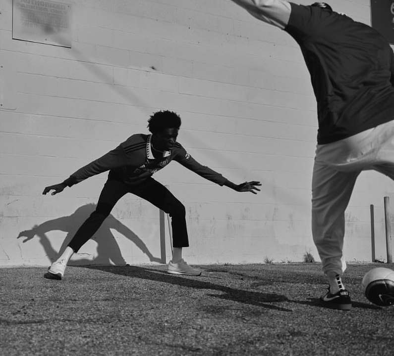 Two athletic men in sportswear play soccer outside on a sunny day, captured in a monochrome medium shot against a textured wall.