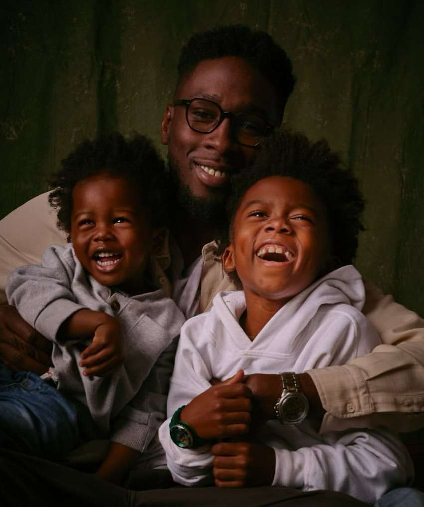 Portrait of a Black father embracing his two young sons, all smiling broadly. The background is a textured green and brown.