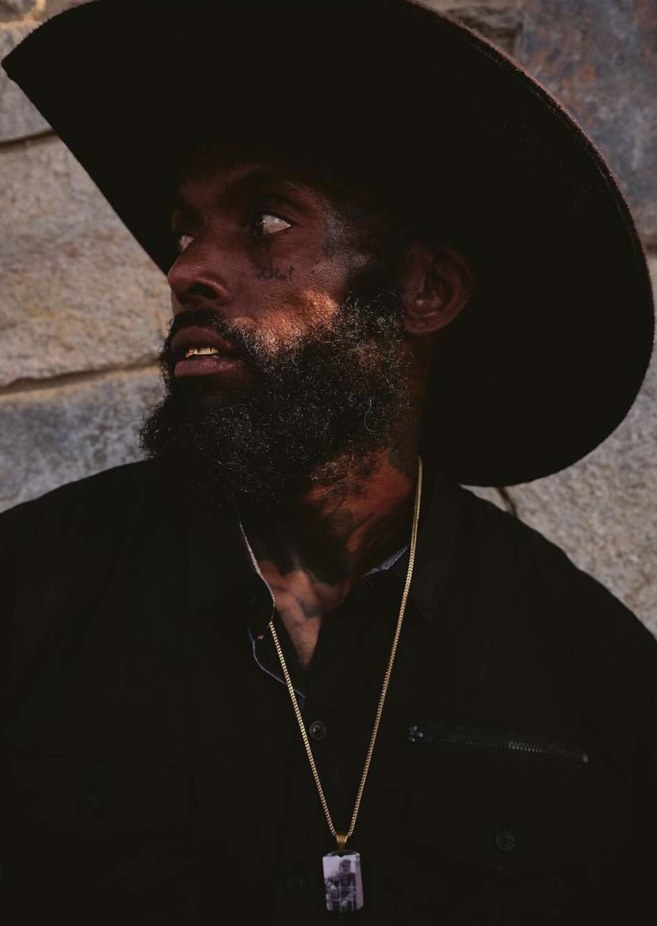 Portrait of an adult male with a beard wearing a black cowboy hat and a gold chain, posing outdoors against a textured stone wall.