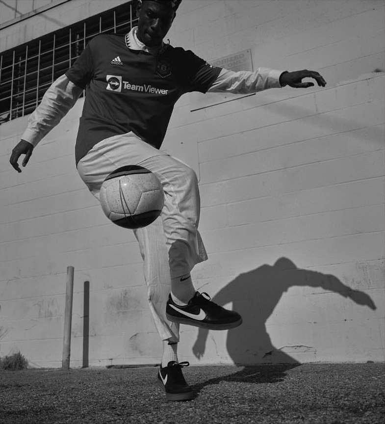 Athletic adult in red soccer jersey balancing a soccer ball on his leg outdoors in front of a textured light grey wall on gravel.