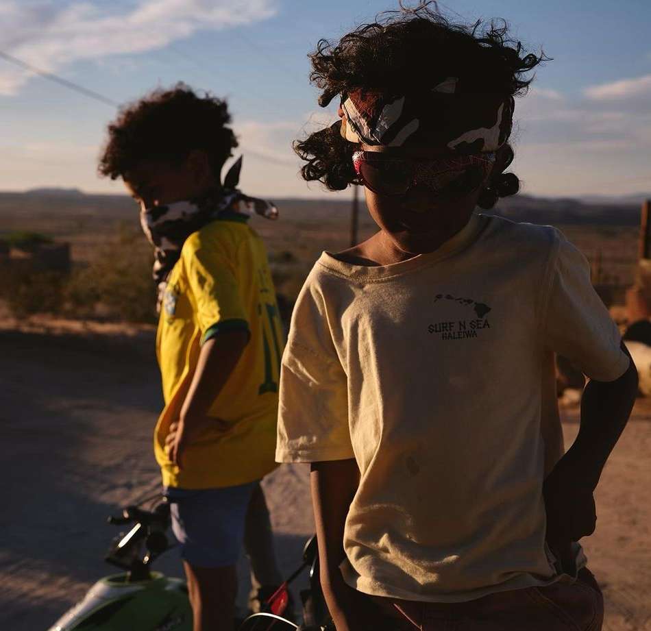 Two children stand outdoors on a sunny day. One child wears a yellow shirt and a patterned bandana. A bike is also visible.