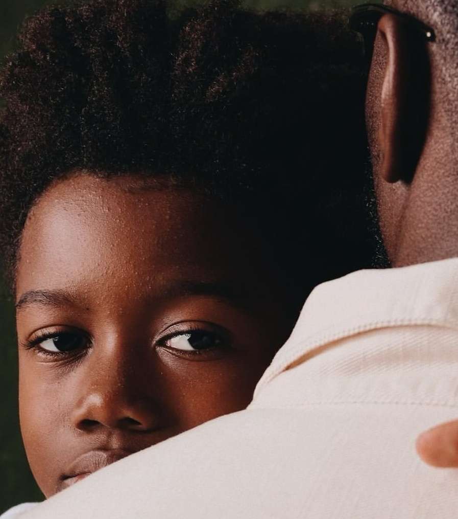 Close-up of a child with dark hair and skin looking directly at the camera, partially obscured by an adult with light skin wearing a light-colored shirt.