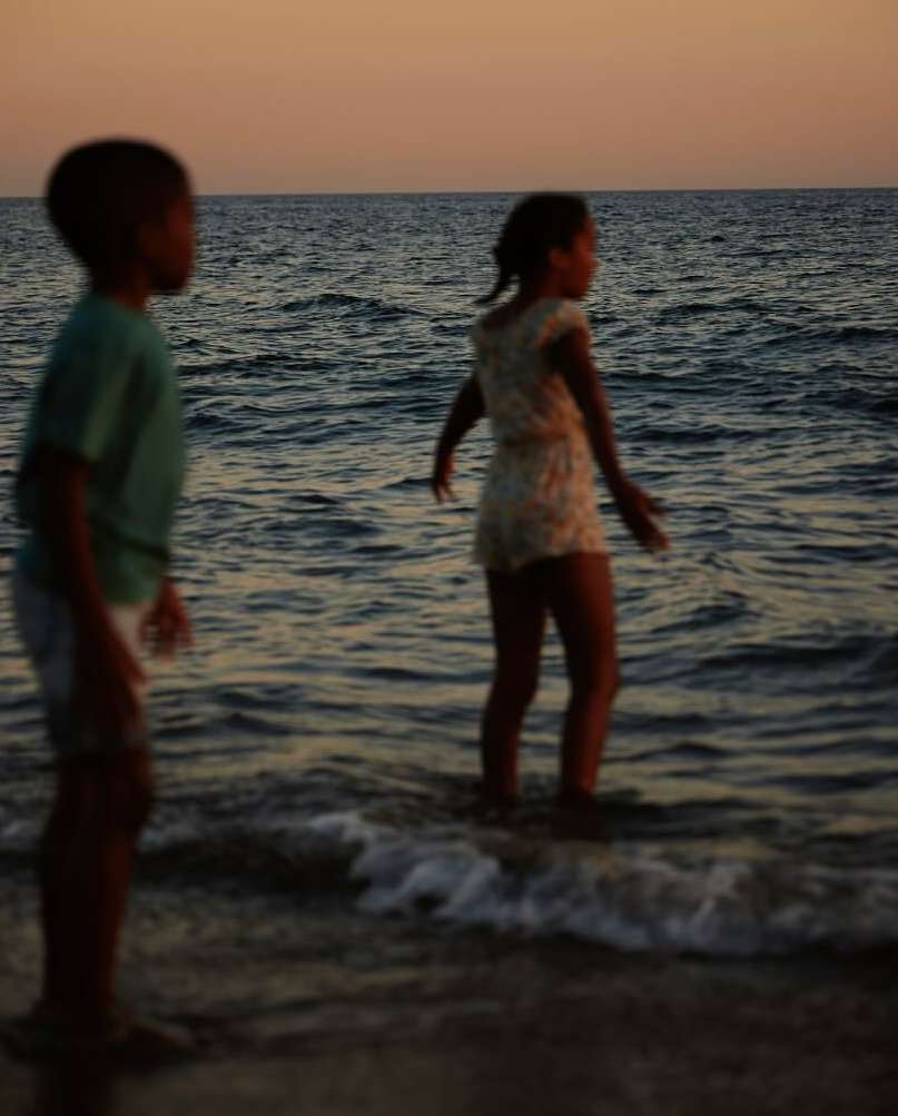 Two kids, a boy and a girl, stand in the ocean at sunset, facing the horizon, with soft light illuminating the water and sky.