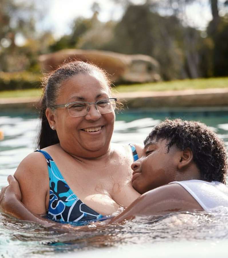 Smiling woman with glasses hugs a boy wearing a white top in a sunlit swimming pool, trees and shrubbery are visible in the background.