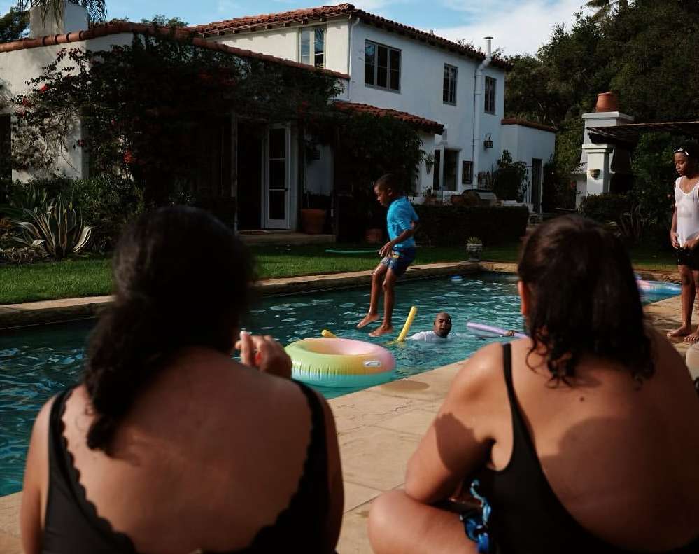 A group of people enjoy an outdoor swimming pool on a sunny day with a child jumping in, houses and green foliage in the background.