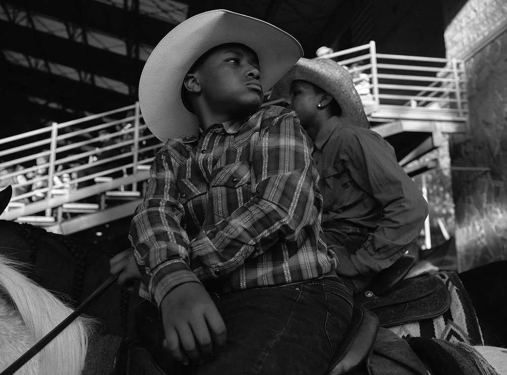 A black and white portrait of two boys on horseback, wearing cowboy hats and shirts, showcasing equestrian style and youthful spirit.
