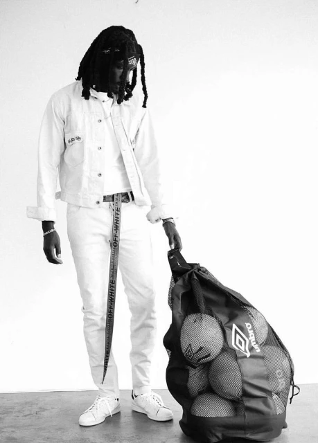 Full body shot of a Black man in all-white casual clothes and long dreadlocks, holding a sports-ball-filled Umbro bag.