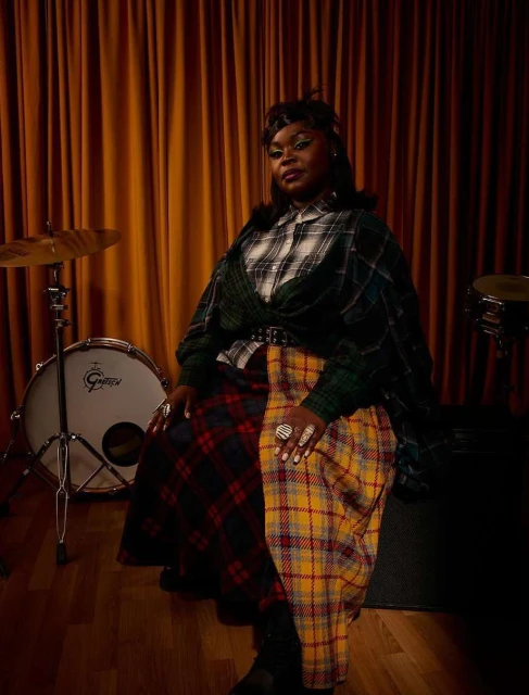 Portrait of an African-American woman in a plaid outfit sitting next to a drum set in a warmly lit interior with brown curtains.