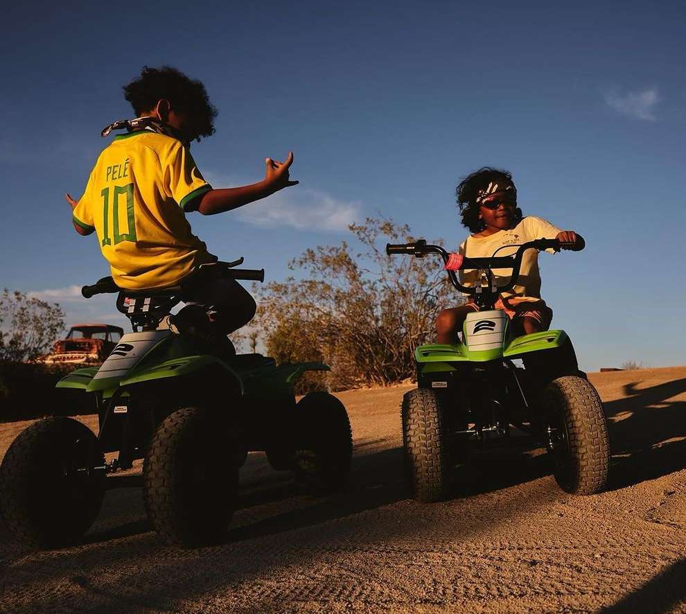 Two children riding green ATVs on a dirt road with desert vegetation under a clear sky, one wearing a Pele soccer jersey.
