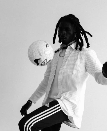 Monochromatic shot of a Black male athlete juggling a soccer ball. He's wearing sportswear and a striped shirt against a light backdrop.