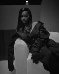 Black and white portrait of a woman in a dark outfit with gloves seated on a white sofa in a studio setting.