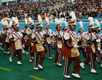Marching band in burgundy and gold uniforms with white plumed shako hats walking across a stadium field.