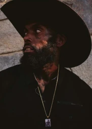 Portrait of a Black man with a beard wearing a wide-brimmed hat and gold chain necklace against stone wall
