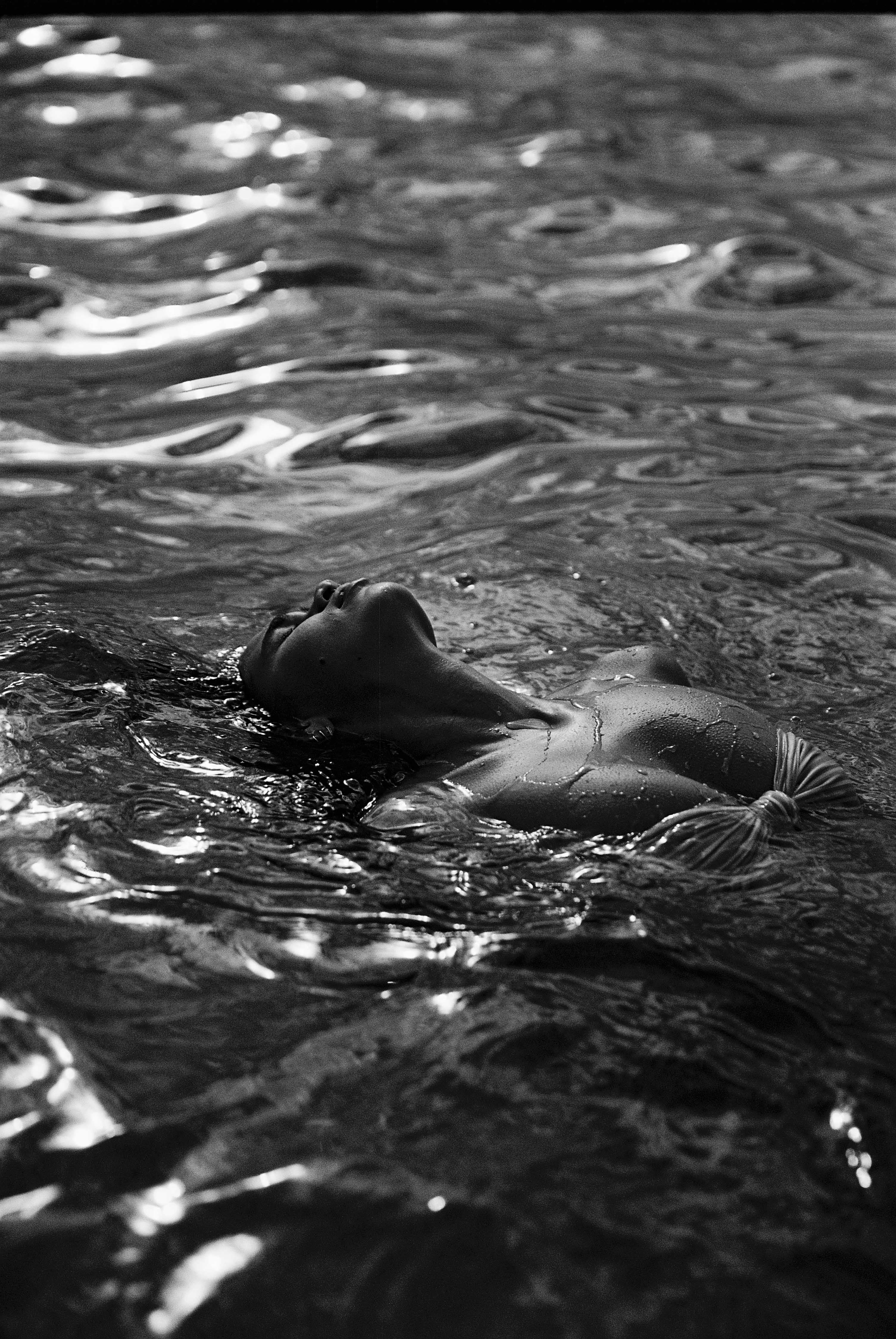 A person floats on their back in water, with their face looking upwards, in a black and white shot.