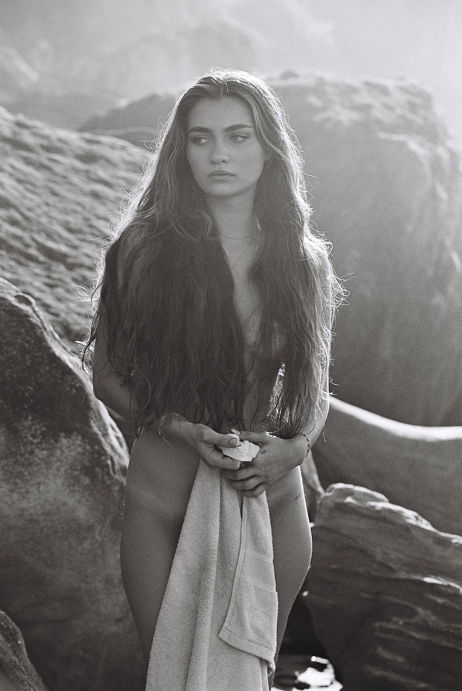 Black and white shot of a young woman standing on a rocky beach holding a towel, long hair flowing down her shoulders.