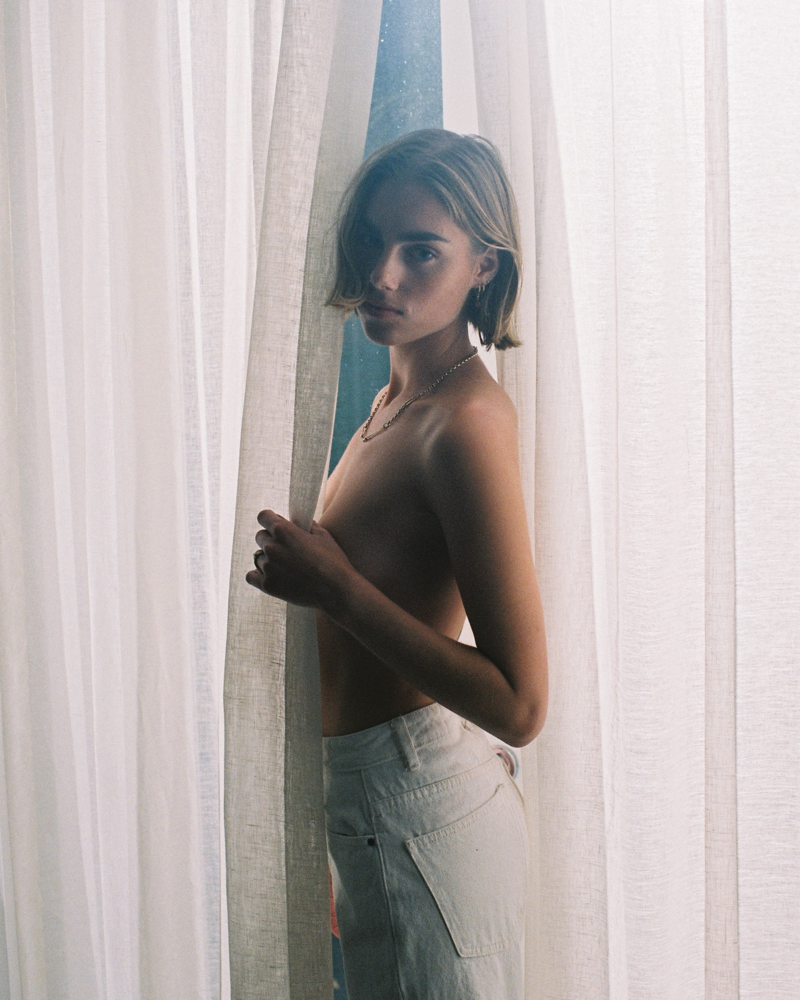 Young woman poses topless wearing jeans, standing in front of light filtering sheer white curtains in a soft, natural light setting.