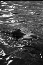 Black and white photograph of a woman floating on her back in dark rippling water, wearing a textured swimwear top.