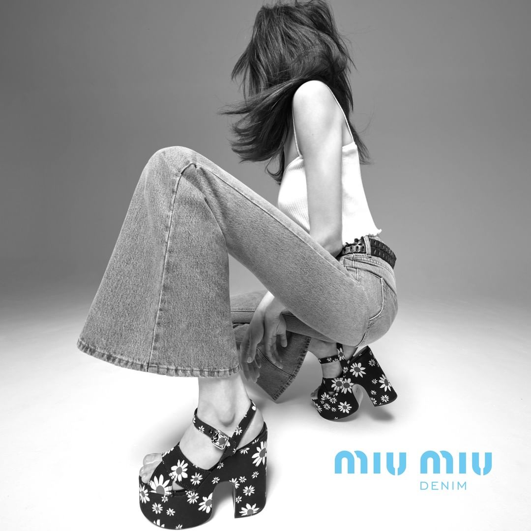 Model in white tank top and flared denim jeans squatting while wearing platform sandals with white daisy patterns.