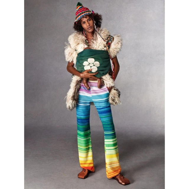 An adult male poses in a photo studio, holding a child in a carrier, wearing striped pants, a hat, fur shawl, and brown leather shoes.