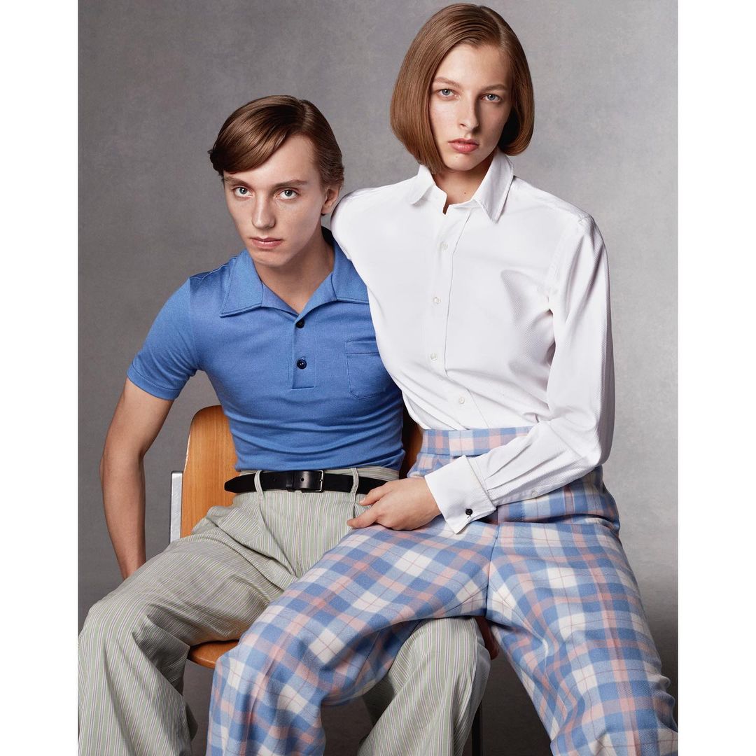 Two fashion models pose indoors against a grey backdrop. The female model sits on the lap of the male model, wearing a white shirt and patterned pants.