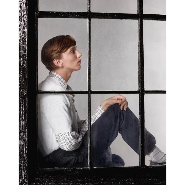 Adult woman poses indoors with serious expression, sitting in a window frame with natural light and a neutral-toned background.