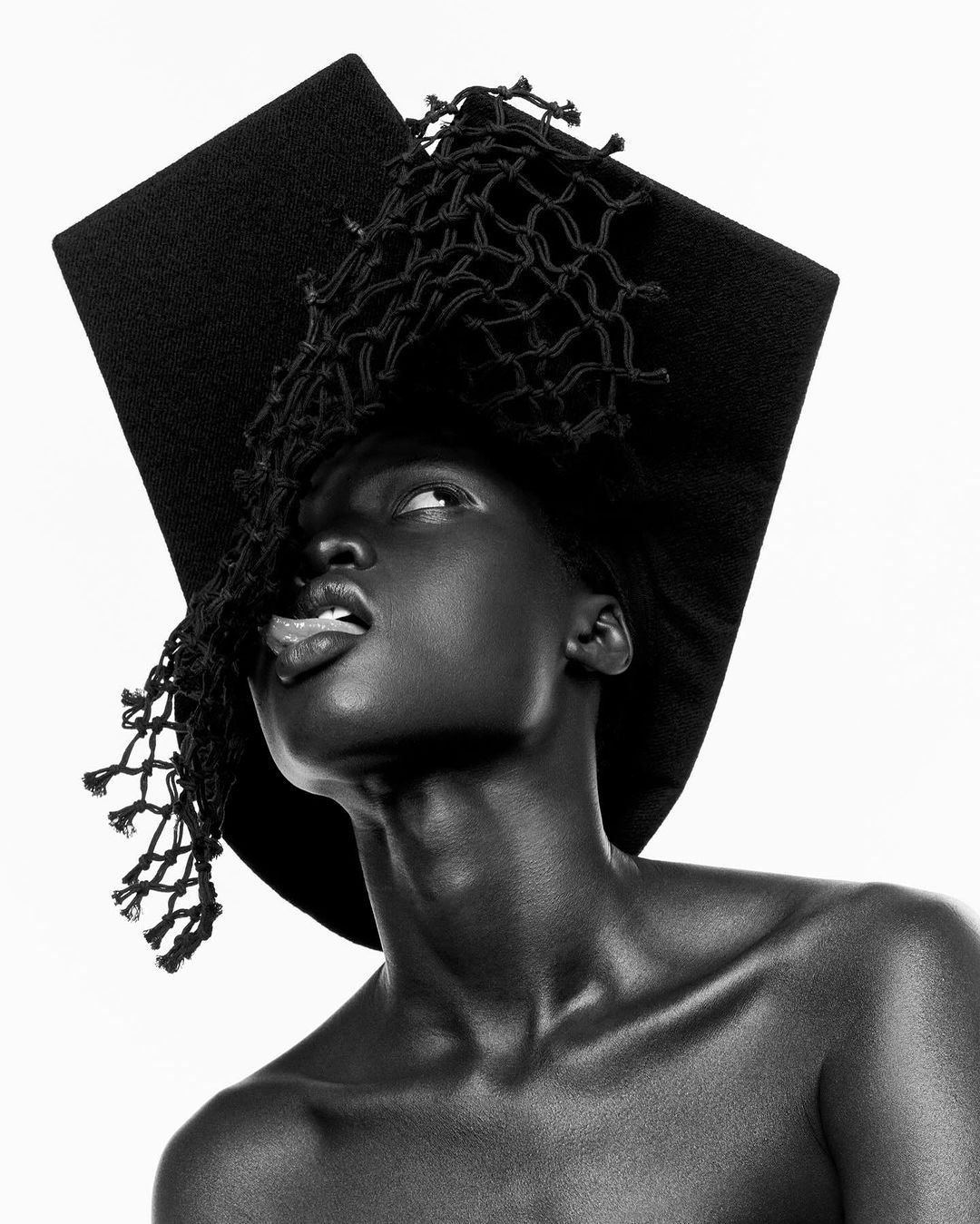 Striking black and white studio portrait of a Black female model wearing a large square hat with net, sticking tongue out, on white.