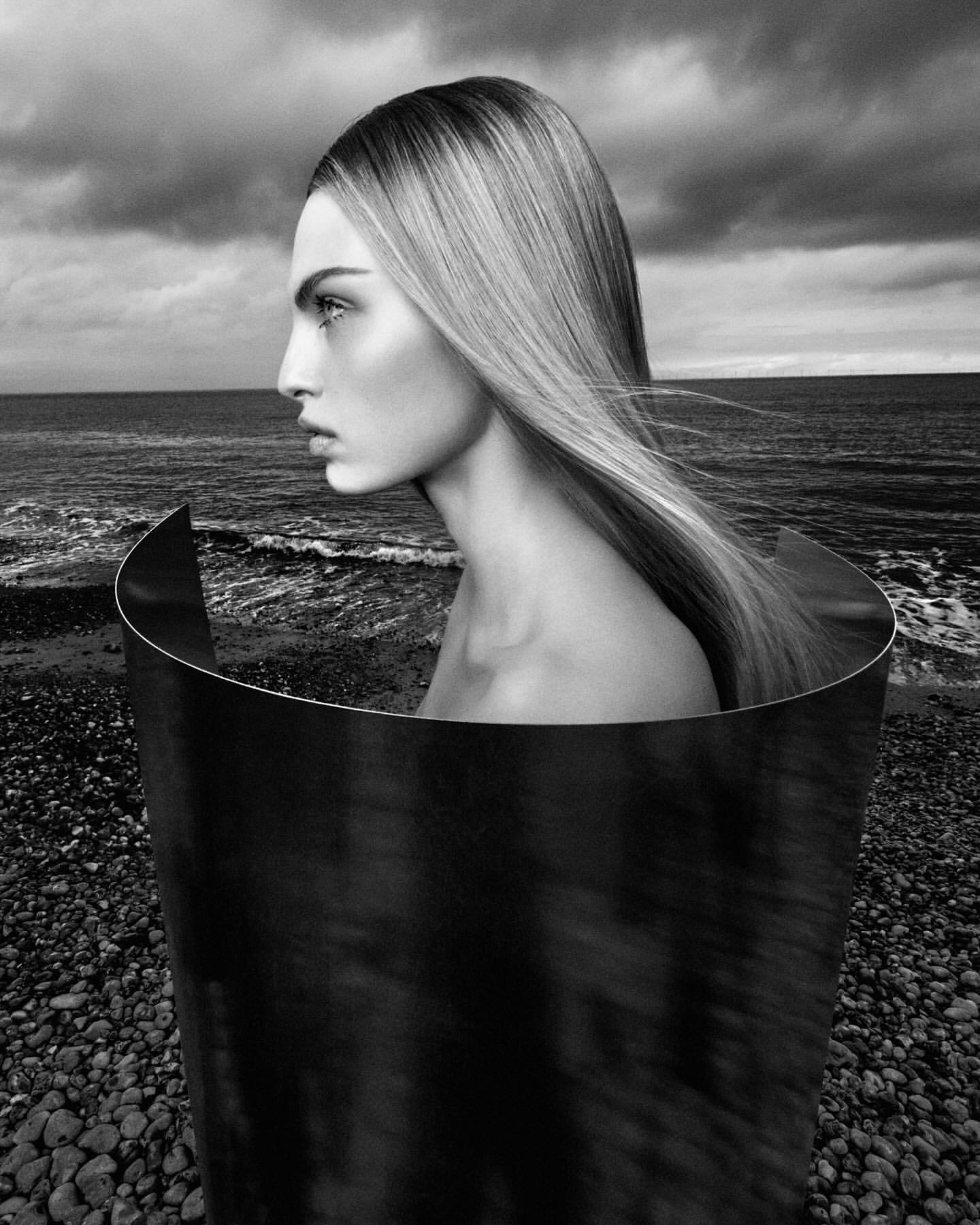 Monochrome portrait of a woman with long blonde hair, captured in profile against a dark ocean and sky within a curved metal frame.