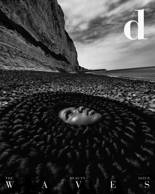 Black and white magazine cover shot of a person's face surrounded by a textured hair pattern against a rocky beach backdrop.