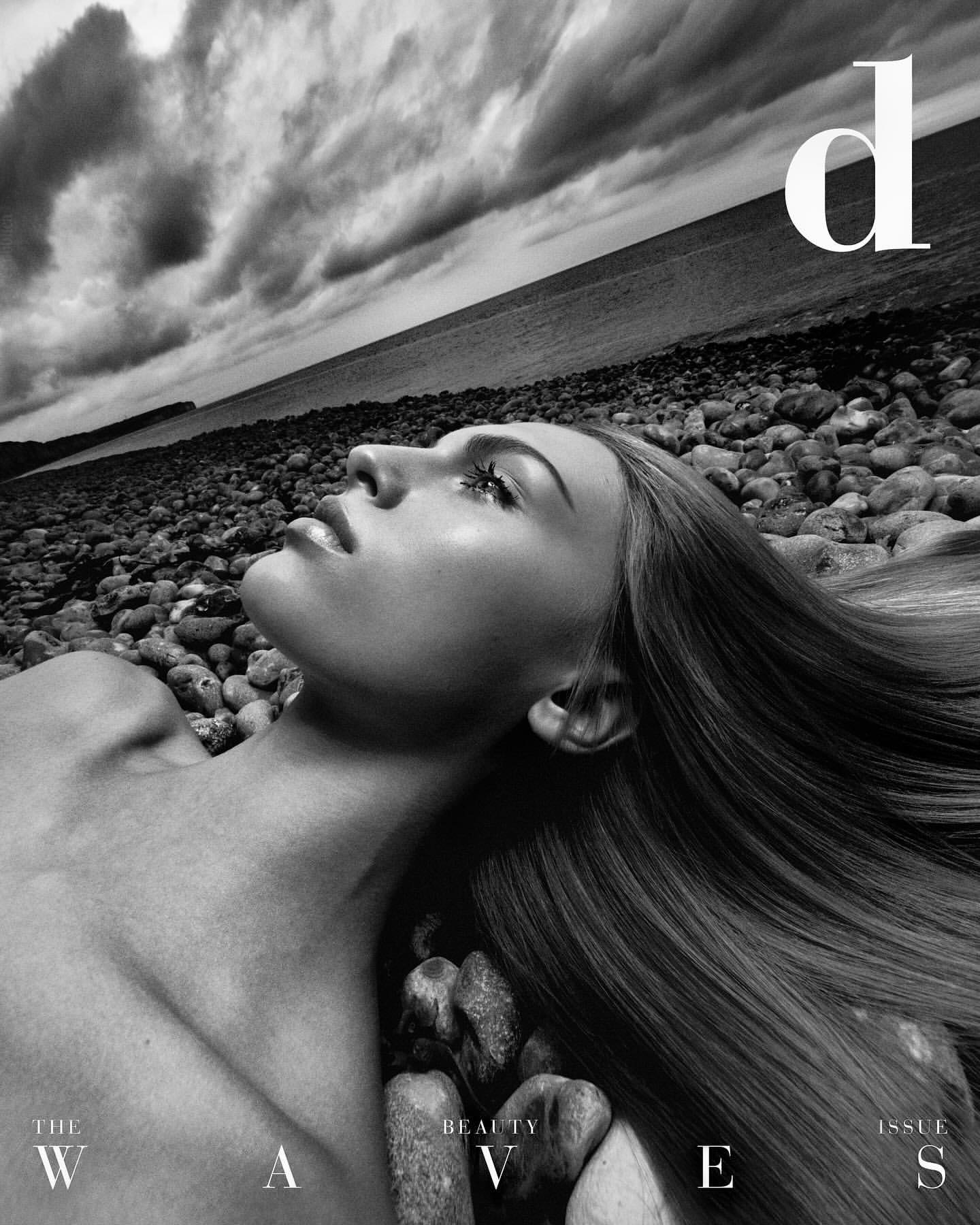 Monochrome beauty shot featuring an adult woman lying on a pebble beach with dramatic sky and ocean backdrop, magazine cover design.