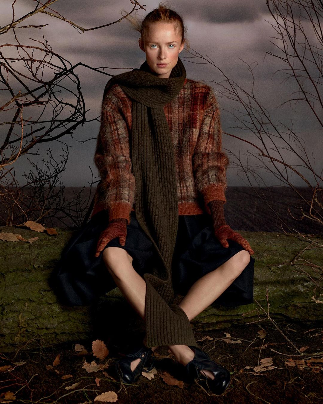 A fair-skinned woman in a patterned sweater and scarf sits cross-legged on a log in a sparse landscape, gazing directly at the camera.