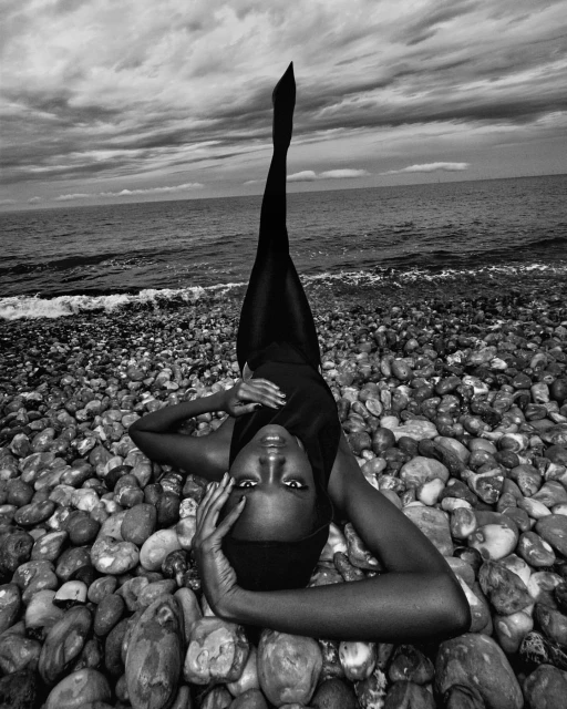 Black and white portrait of an adult woman in a bodysuit laying on a pebble beach with legs raised to the sky.