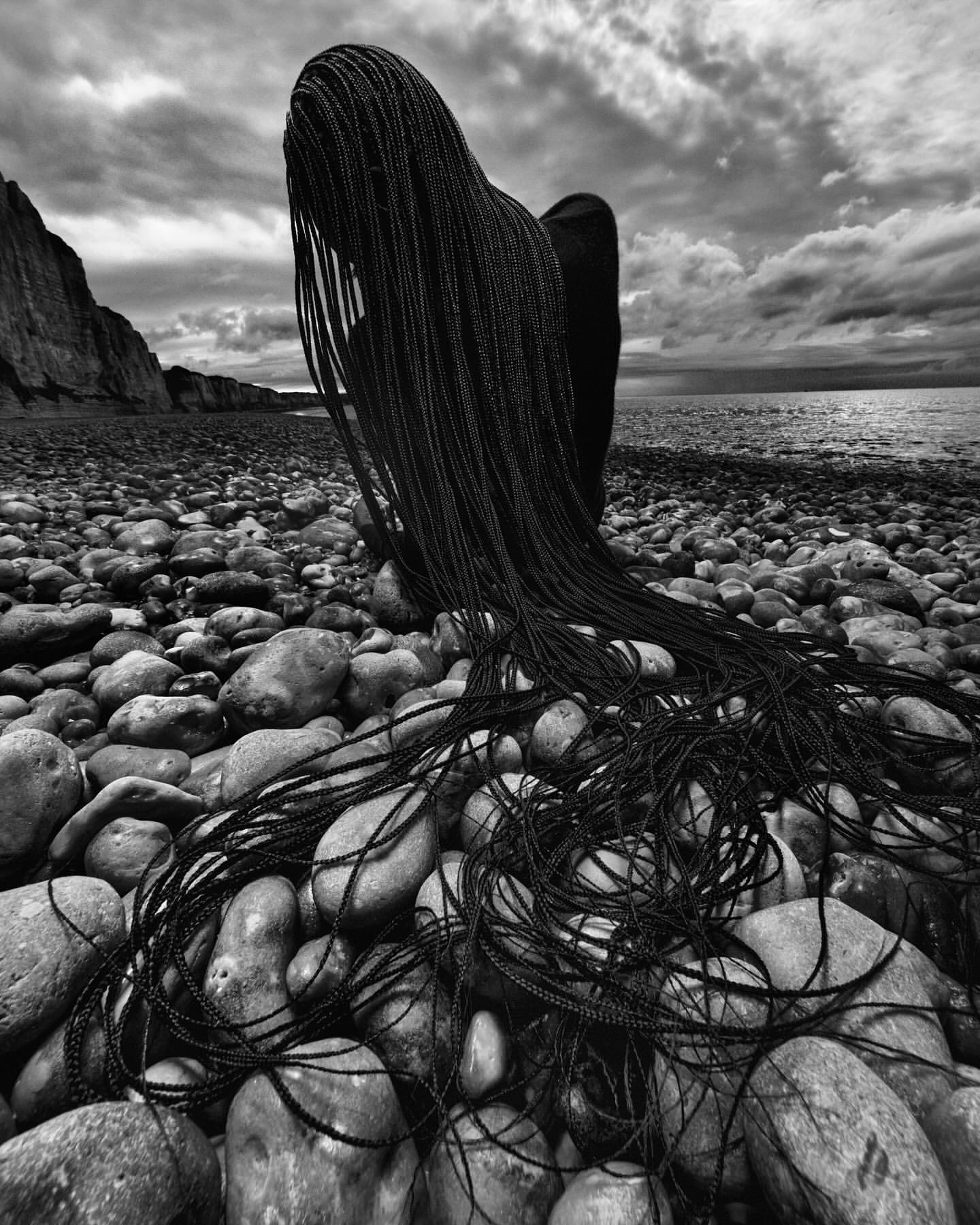 Monochrome portrait of a person with long braided hair seated on a pebble beach with rocky cliffs and cloudy sky in the background.