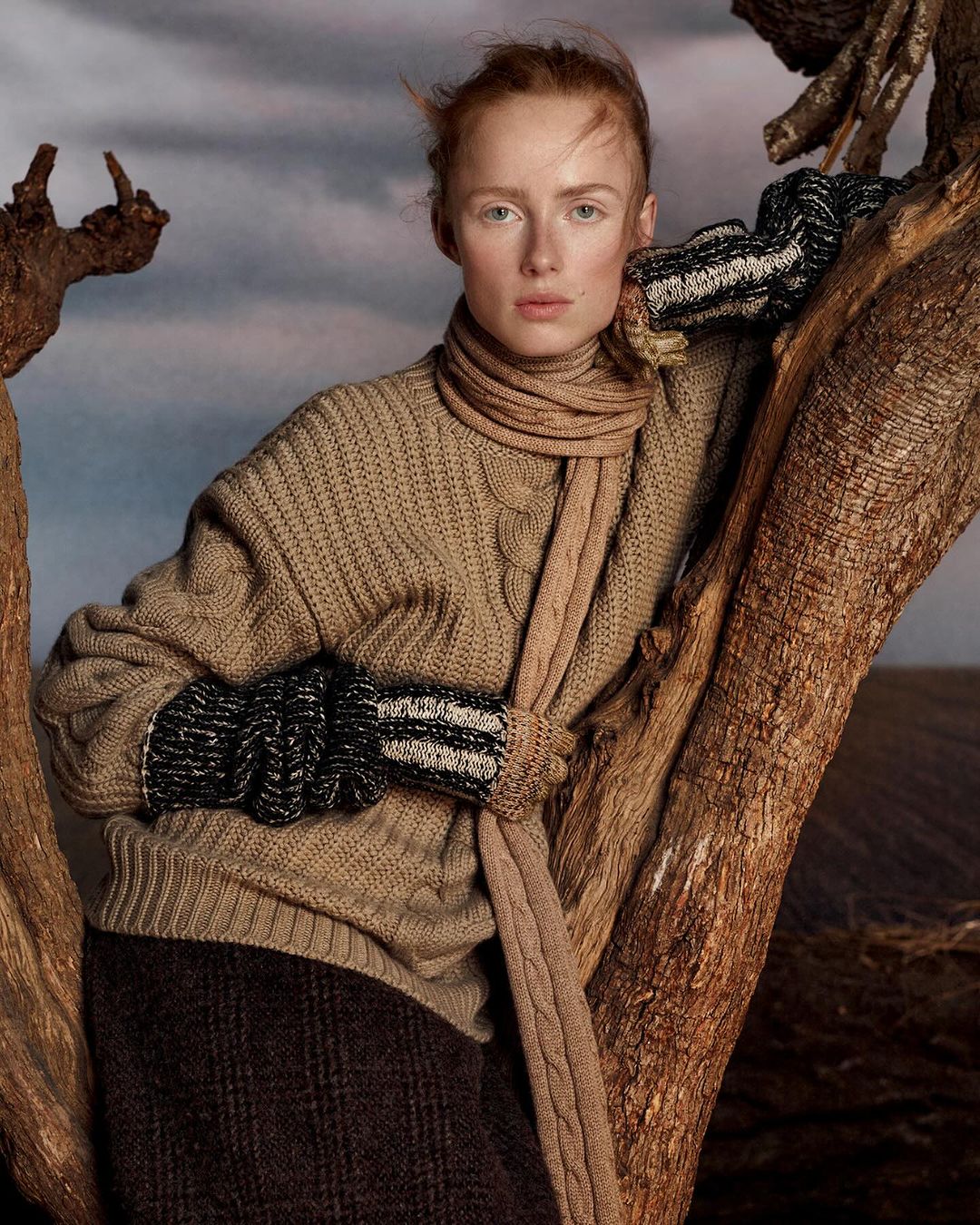A redhead model wearing a tan sweater and scarf poses near a tree in a field under a cloudy sky for a fashion editorial shot.
