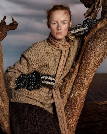 A redhead model wearing a tan sweater and scarf poses near a tree in a field under a cloudy sky for a fashion editorial shot.