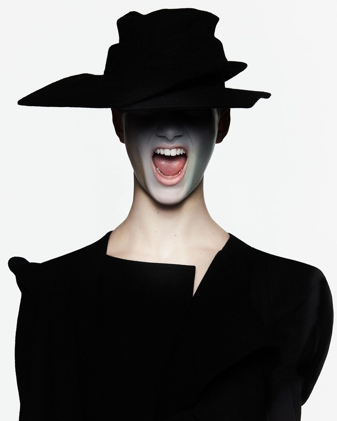 An adult female with face paint and wearing a sculptural hat and black garment in a studio setting with neutral backdrop.