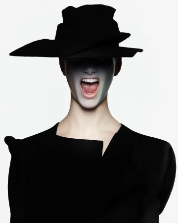 An adult female with face paint and wearing a sculptural hat and black garment in a studio setting with neutral backdrop.