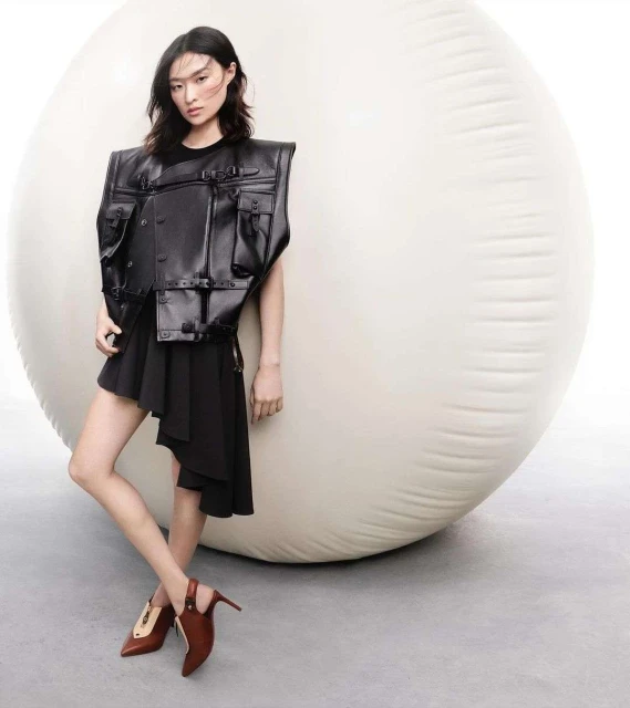 A stylish Asian woman poses against a large white ball, wearing a black vest, dress, and brown heels, in a studio setting with grey flooring.