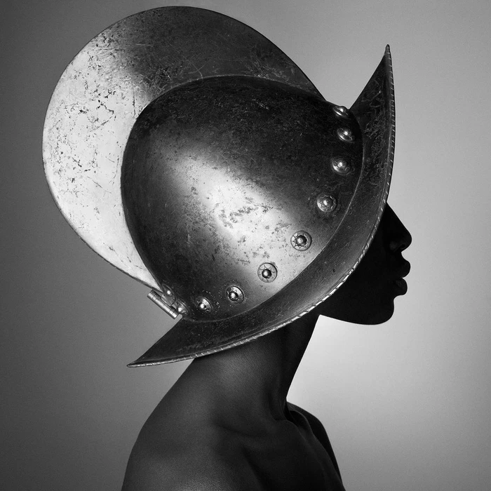 Profile of a person wearing an elaborate dark metallic helmet with a large circular crest, photographed by Julien Martinez Leclerc.