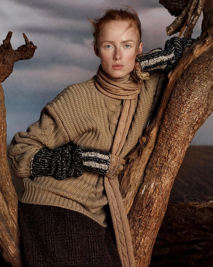 Redheaded woman in a textured camel sweater leaning against a gnarled tree branch under an overcast sky.