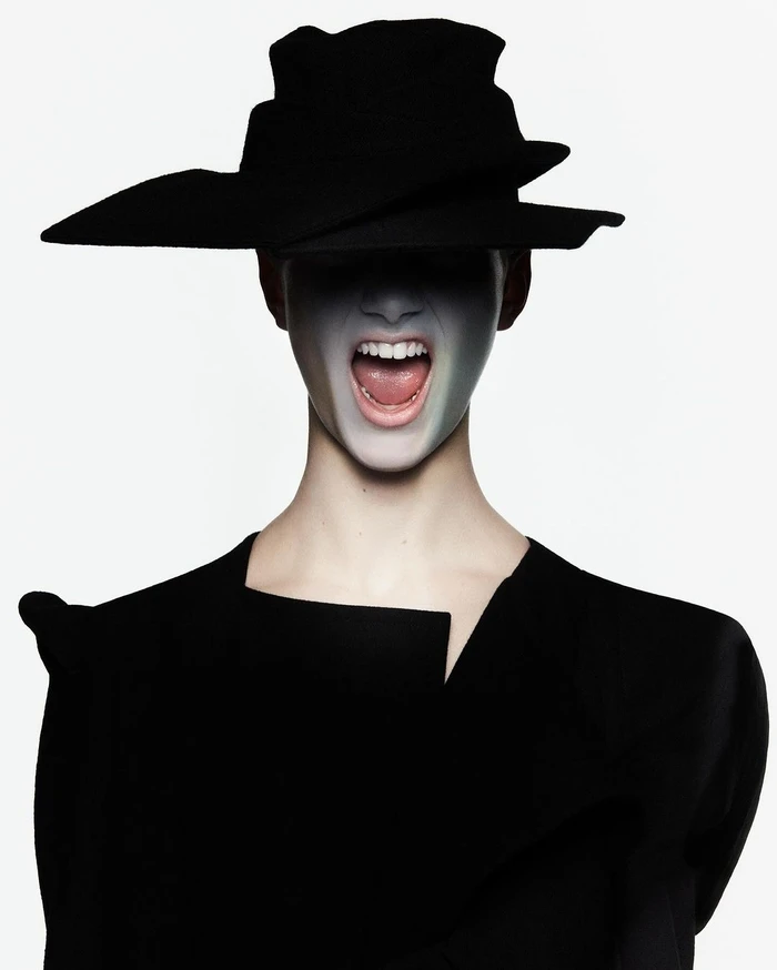 Model wearing a structured black hat and jacket with white painted face and open mouth on a solid white background